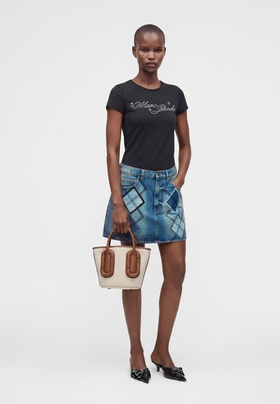 Γυναίκα που φοράει μαύρο t-shirt με το κείμενο "Marc Jacobs", τζιν φούστα με σχέδια patchwork, μαύρες μυτερές γόβες και κρατάει μια μπεζ και καφέ τσάντα.