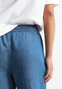 Denimové kalhoty s elastickým pasem, zobrazené zezadu. Mají středně modrou barvu a hladký povrch, doplněné o bílé tričko.