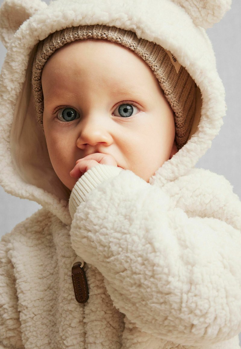 Hvid fleece overtøj med en tekstureret overflade, der har bjørne-ører på hætten, en ribbet beige strikhue og et lædermærke.