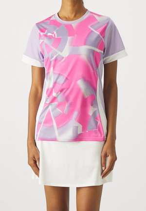 Vrouw draagt een sportshirt met korte mouwen in roze en lavendel met een abstract patroon, gecombineerd met een witte rok tegen een effen achtergrond.