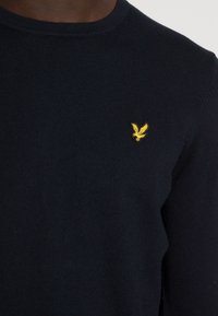 Maglione lavorato a maglia in navy con collo a giro, presenta un piccolo logo di un'aquila gialla sul petto. Tessuto liscio, design aderente.