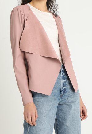 Woman wearing light pink open-front jacket with wide lapels over white shirt and high-waisted light blue jeans, standing against a plain background.