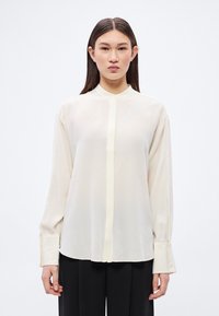 Fiatal nő egyenes, sötét hajjal laza, hosszú ujjú krémszínű blúzt visel mandzsettagallérral és fekete nadrágot, fehér háttér előtt áll.