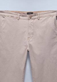Pantaloni in cotone beige con zip, chiusura a bottone e passanti per cintura. Presentano tasche anteriori oblique e dettagli di cucitura discreti.