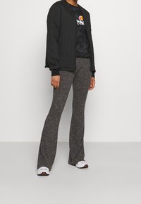 Persona vistiendo una chaqueta acolchada negra, una camiseta gráfica negra, pantalones de punto gris de campana y zapatillas blancas frente a un fondo sencillo.