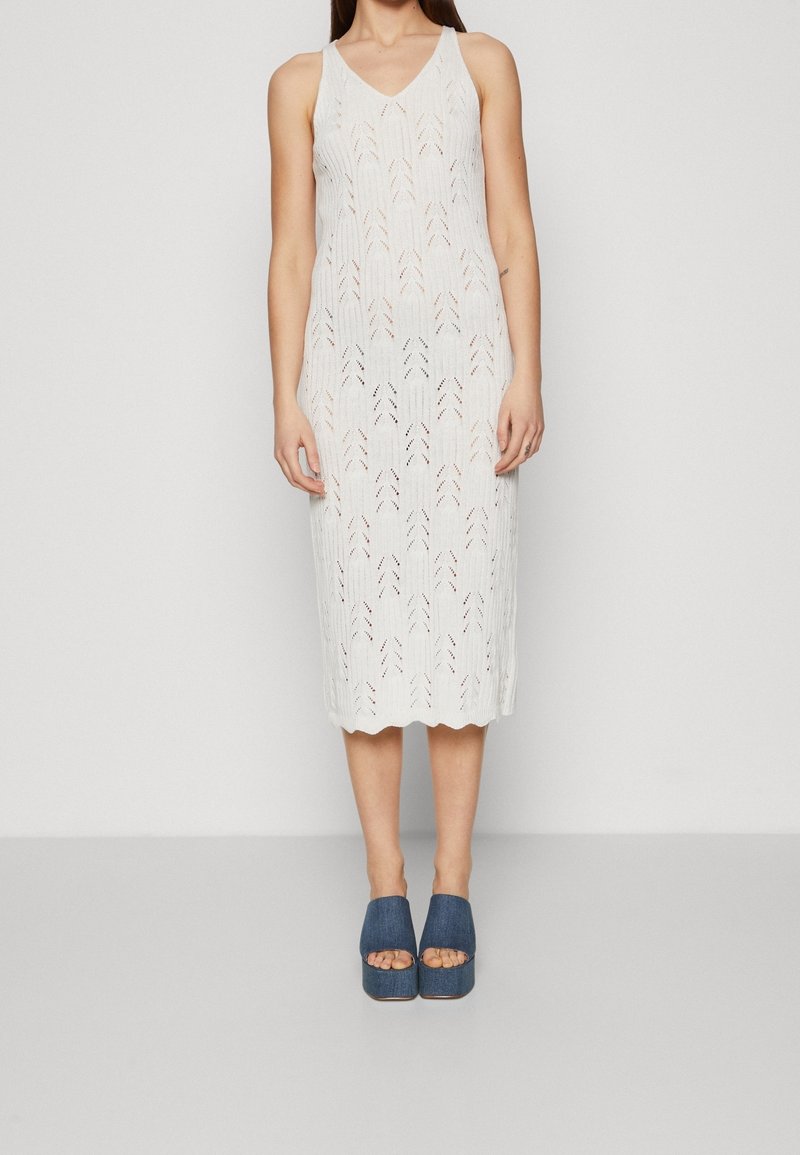 Femme portant une robe midi tricotée blanche sans manches avec un col en V et des chaussures à plateforme bleues ouvertes à l'avant, debout devant un fond uni.