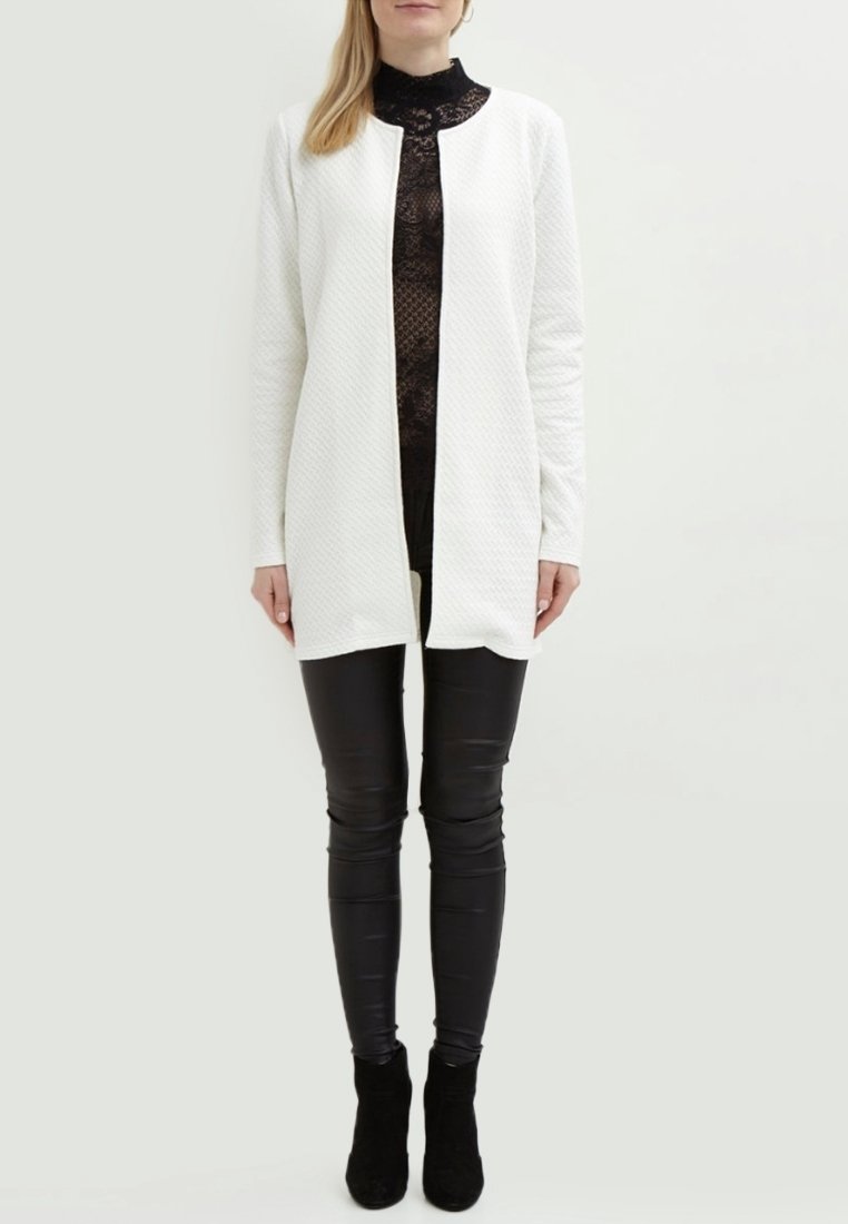 Femme portant un long cardigan blanc texturé ouvert sur le devant, un top en dentelle noire, un pantalon en cuir noir et des bottines noires, debout devant un fond uni.