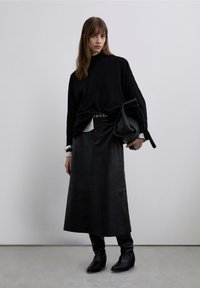 Svart stickad tröja, svart lädermidi-kjol, tajta svarta stövlar; lager på lager av vit skjortmanschett synlig, överdimensionerad svart väska i handen.