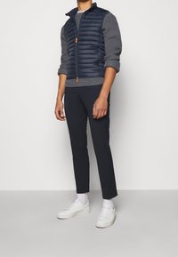 Navy puffer vest with a zip front over a grey knit sweater, paired with dark trousers and white sneakers. Textured fabrics, minimal design.