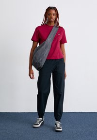 Rotes Baumwoll-T-Shirt, graue Umhängetasche, schwarze tapered Hose und schwarze Sneakers mit weißen und grauen Akzenten auf einem blauen Teppichhintergrund.