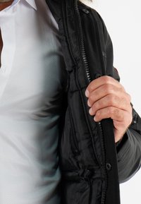 Veste noire avec une fermeture éclair à l'avant et doublure en tissu. Une main tient la tirette de la fermeture éclair, révélant une couche interne à texture plus claire et une poche.