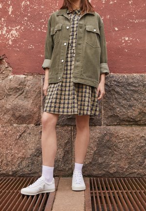 Olivově zelená kordurová bunda přes černou a žlutou károvanou šaty, doplněná bílými teniskami a ponožkami do půli lýtek, stojící na kovové mřížce.