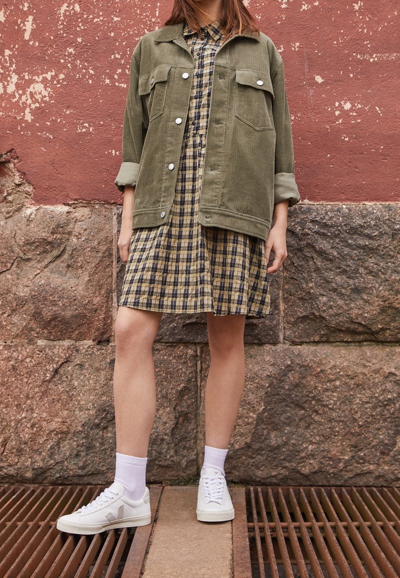 Olivově zelená kordurová bunda přes černou a žlutou károvanou šaty, doplněná bílými teniskami a ponožkami do půli lýtek, stojící na kovové mřížce.