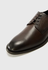 Zapato de vestir de cuero marrón con punta lisa, parte superior texturizada con cordones y una sutil costura en contraste a lo largo de la suela.