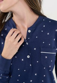 Camiseta de manga larga azul marino con patrón de estrellas blancas, cuello redondo, cierre de botones en la parte delantera y bolsillo con detalle de ribete blanco. Textura de tela suave.