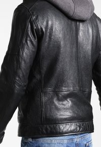 Veste en cuir noir avec une texture lisse, un design structuré et une capuche. Détails de couture visibles et deux poches latérales sont présents.