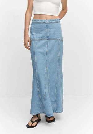 Femme portant une longue jupe en denim bleu clair, un haut court blanc sans manches et des sandales plates noires, debout sur un fond blanc uni.