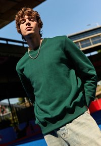 Groene crewneck sweatshirt in een effen ontwerp, met een geribbelde halslijn en manchetten. Gecombineerd met lichtblauwe beschadigde jeans.