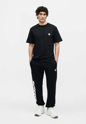 Jeune homme debout les mains dans les poches, portant un t-shirt noir et un pantalon de survêtement avec des logos smiley jaunes et des baskets blanches.