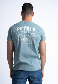Lichtblauwe t-shirt met korte mouwen en een grafische print van surf, palmbomen en de tekst "PETROL WAVERIDERS." Zachte katoentextuur.
