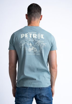 T-shirt à manches courtes bleu clair avec un imprimé graphique représentant des surf, des palmiers et le texte "PETROL WAVERIDERS". Texture en coton doux.