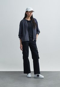 Ciemnoszara kurtka typu bomber na zamek, założona na czarną koszulkę, w połączeniu z czarnymi spodniami cargo i białymi sneakersami. Czapka dodaje swobodnego charakteru.