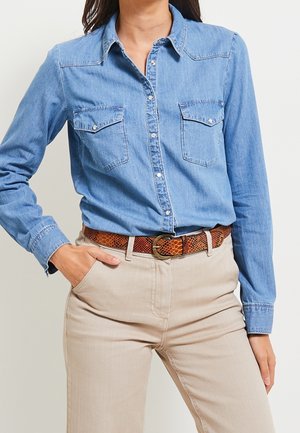 Vrouw draagt een blauw denimoverhemd met knopen, ingestopt in beige broeken met een bruine gestructureerde riem, één hand in de zak.