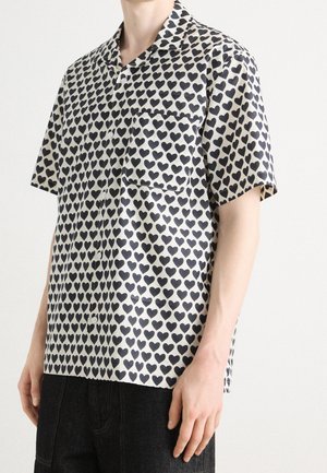 Mężczyzna w koszuli na krótki rękaw z czarnym wzorem serc i kieszenią na piersi, w ciemnych spodniach na jednolitym tle.