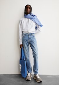 Gestreepte blauw-witte overhemd, lichtblauwe jeans, grijze sneakers en een blauwe totebag met een grote witte letter. Textuurdetails van de stof zichtbaar.