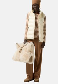 Krémová prošívaná vesta s kapucí, hnědé a bílé pruhované tričko s dlouhým rukávem, hnědé kalhoty s širokými nohavicemi a načechraná béžová kabelka. Jemné textury po celém outfitu.