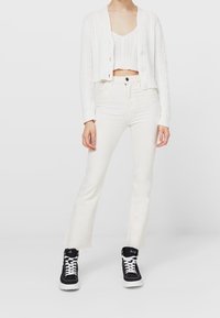 Biała kardigan z splotem w warkocze, biały top na ramiączkach, białe jeansy dzwony, w zestawieniu z czarnymi sneakersami za kostkę z białymi sznurowadłami.