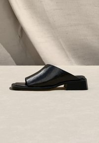 Μαύρο mule σανδάλι ανοιχτής μύτης με χαμηλό τακούνι και φαρδιά, γυαλιστερή δερμάτινη λουρίδα στο πόδι, σε ουδέτερο υφασμάτινο φόντο.