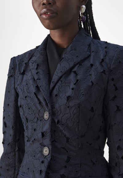 Navy blue lace blazer featuring a floral pattern, structured silhouette, and two dark buttons. The fabric has a textured, layered appearance.