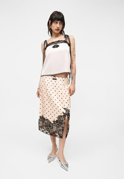 Γυναίκα φοράει λευκή αμάνικη μπλούζα με μαύρη δαντέλα στο τελείωμα και μπεζ midi φούστα με μαύρες πουά και δαντελένιες λεπτομέρειες, ασημένια μυτερά παπούτσια.