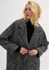 Kabát s klasickým vzorom houndstooth v čierno-bielej farbe so širokým golierom, oversized strihom a veľkými gombíkmi, s textúrovanou tkaninou a dlhými rukávmi.