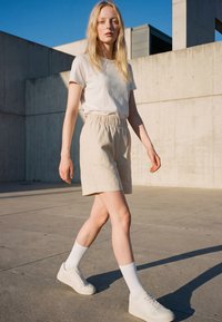 Junge Frau, die draußen in beigen Shorts, weißem T-Shirt, weißen Socken und weißen Turnschuhen vor einem Betongebäude geht.