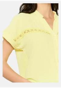 Camicia gialla chiara con maniche corte arrotolate, chiusura con bottoni e dettagli in pizzo sulla parte superiore del petto. Tessuto testurizzato.