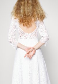 Robe blanche en dentelle avec des manches longues, un décolleté profond au dos et une jupe fluide, ornée d'un motif en dentelle détaillé et de bords de manches à franges.