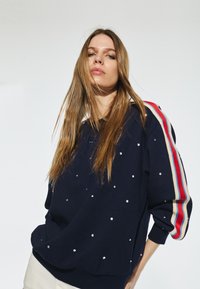 Marinblå sweatshirt med rundad nederkant, prydd med silvernitar, med röd, vit och beige randiga detaljer på ärmarna.