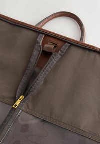 Mânerele din piele maro sunt atașate la o geantă din material gri, care prezintă un fermoar aurii, cu o textură netedă și elegantă și un model subtil pe suprafață.