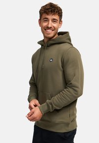 Lächelnder Mann mit lockigem Haar, der einen olivgrünen Hoodie mit kleinem Logo auf der Brust trägt und vor einem einfarbigen Hintergrund steht.