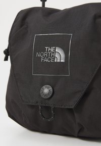 Fekete szövet táska, hajtogatott fedéllel, kör alakú fekete gombbal és szürke "THE NORTH FACE" logóval. Texturált felület finom vonalakkal.