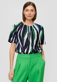 Blouse à manches courtes en bleu marine avec des rayures abstraites vertes et blanches, dotée d'un col rond et d'une coupe ample. Associée à un pantalon vert taille haute.
