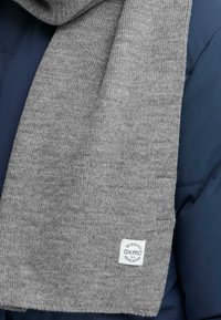 Grå strikket tørklæde med hvid Oxmo-label, der hænger over en navy blå polstret jakke.