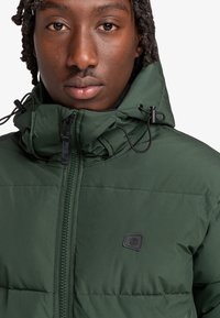 Giacca imbottita verde con colletto alto, zip frontale e toggle regolabili. Include una piccola patch nera con logo sul petto.