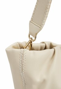 Borsa a tracolla in pelle beige con superficie texturizzata, hardware dorato e dettagli cuciti lungo il lato. Parte superiore aperta con una forma morbida.