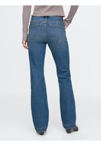 Kvinna står med avslappnade händer, iklädd högmidjade blå denimjeans, beige långärmad tröja och svarta klackskor mot en vit bakgrund.
