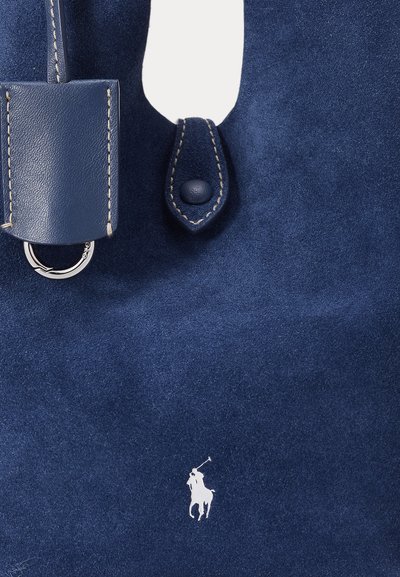 Marinblå mockaväska med texturerad yta, läderdetaljer, en framträdande silverfärgad hårdvarurring och en vit logotyp av en polospelare.