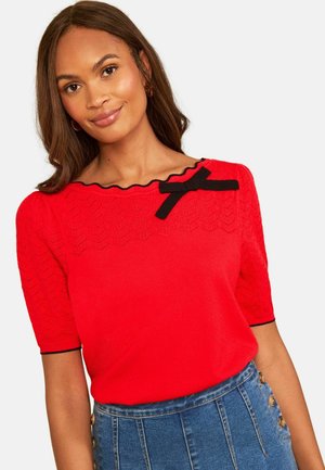 Kvinde iført en rød T-shirt med korte ærmer med sort kant og sløjfe ved halsudskæringen, kombineret med en højtaljet blå denim nederdel med knapper.