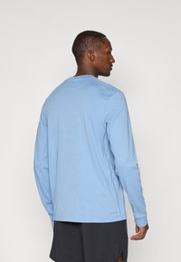 Šviesiai mėlyna ilgos rankovės marškinėlių su minkštos medvilnės mišiniu, su apvalia iškirpte ir šoninėmis siūlėmis, vilkima su tamsiomis šortais.